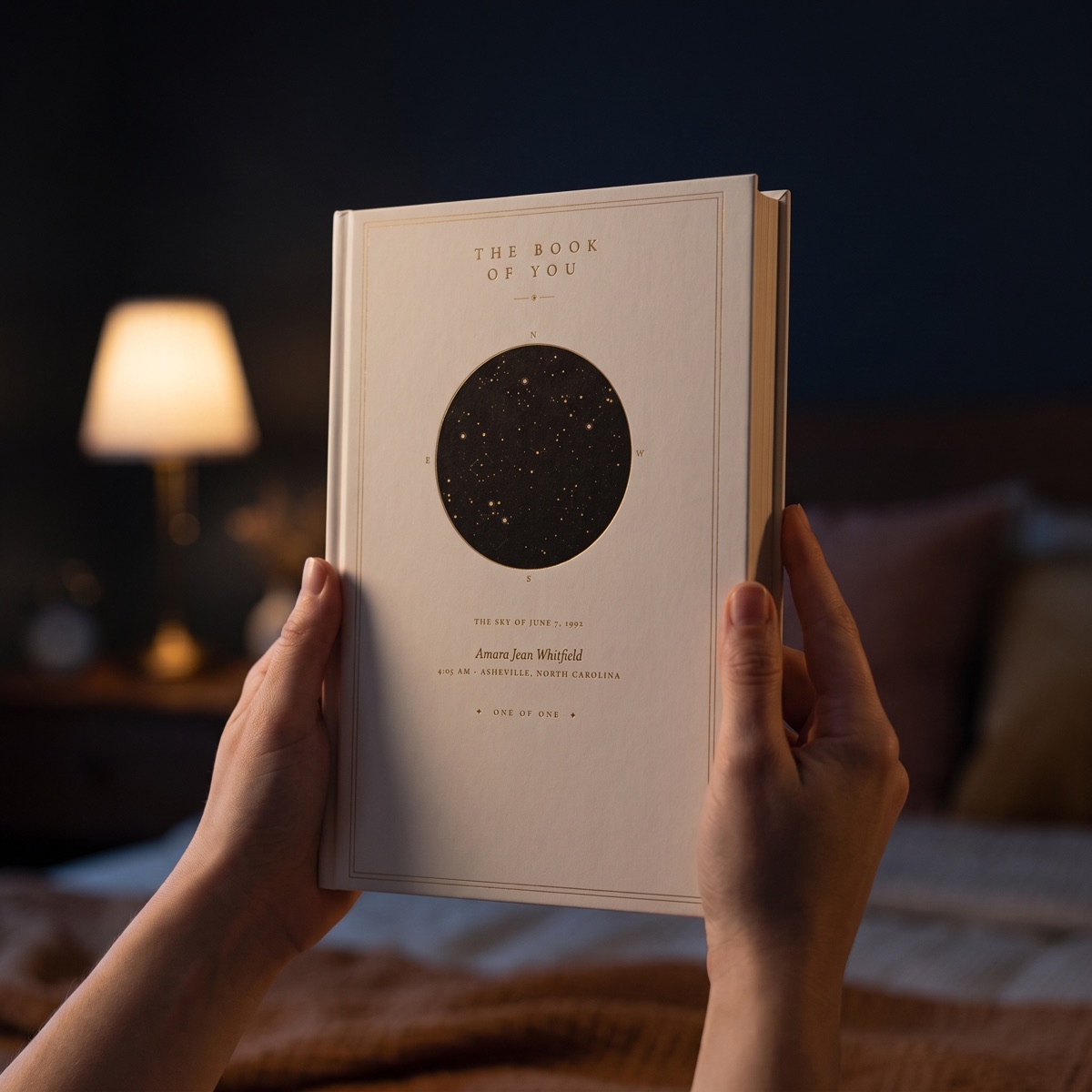 The Book of You held in two hands by lamplight — a matte hardcover, the birth sky in gold on a white board